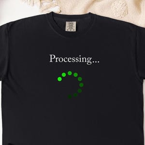 May include: Black t-shirt with the text "Processing..." above a green loading circle graphic. The shirt is a solid black color and appears to be made of a soft material. The design is centered on the front of the shirt.