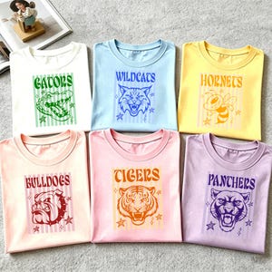 May include: Six folded t-shirts in various colors, each featuring a team mascot and name. The shirts are white, light blue, yellow, pink, and lavender. The graphics include a gator, wildcat, hornet, bulldog, tiger, and panther.
