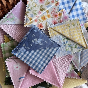 May include: A collection of square fabric bookmarks with pinked edges, featuring various patterns including blue and white gingham, floral prints, yellow checkered designs, and textured pink fabric, all layered together in a woven basket.