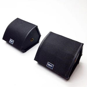 Puede incluir: Dos estuches negros, curvos y rectangulares con una superficie texturizada. Cada estuche tiene una pequeña etiqueta rectangular con la palabra "TACT" en blanco. Un estuche tiene una arandela de color latón. Los estuches están sobre un fondo blanco.