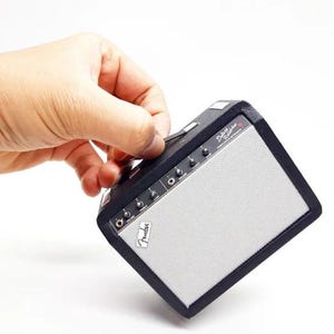 Puede incluir: Un amplificador Fender Deluxe Reverb en miniatura, negro y plateado. El amplificador tiene una cubierta de altavoz plateada, un panel de control negro con perillas y el logotipo de Fender. El amplificador está sostenido por una mano.