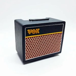 Puede incluir: Un amplificador de guitarra negro en miniatura con una cubierta de altavoz de malla marrón y la palabra "VOX" en letras doradas. El amplificador tiene un asa negra en la parte superior y una franja amarilla. Es una pequeña réplica detallada.