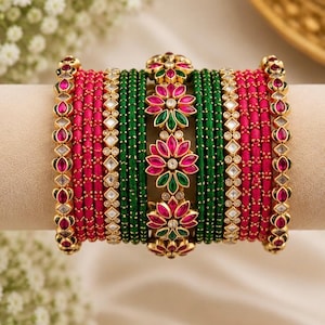 Pode incluir: Um conjunto de pulseiras empilhadas com faixas alternadas de fio rosa vibrante e verde escuro. O conjunto inclui pulseiras de metal dourado com pedras transparentes incrustadas e uma fileira central de ornamentos florais com pétalas rosas e verdes.
