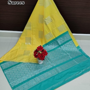 May include: A bright yellow saree featuring silver square patterns and a wide teal border with intricate silver floral motifs. The text Soft Silk Sarees is visible in the upper left corner. A small vase with red flowers sits on the fabric.