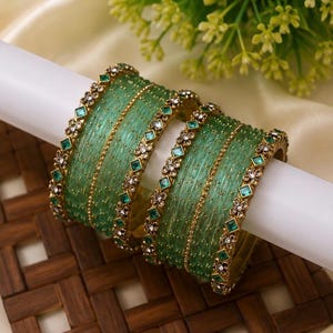 Pode incluir: Um conjunto de pulseiras verde translúcido com superfície texturizada e bordas de metal dourado. As pulseiras são decoradas com pequenas pedras verdes de corte quadrado e cristais claros brilhantes em um padrão floral repetitivo.