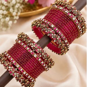 Puede incluir: Un conjunto de brazaletes de color magenta intenso con una superficie texturizada y detalles florales metálicos. Cada brazalete está adornado con pequeñas piedras transparentes y brillantes en forma de flor sobre una varilla de soporte.
