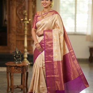 May include: A cream-colored saree featuring a wide, deep magenta border with intricate gold zari woven patterns. The fabric has a subtle vertical striped texture, and the garment is draped with a pleated front section and a long pallu draped over one shoulder.