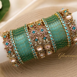 May include: A set of bangles featuring translucent teal glass bands interspersed with gold-tone metal spacers. The set includes decorative floral-shaped links adorned with small pink and teal crystals. The text Glowvanadesigns is visible.