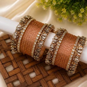 Pode incluir: Um conjunto de dois braceletes cor de cobre com superfície texturizada e bordas metálicas ornamentadas. Cada bracelete é acentuado com pedras transparentes em forma de marquise num padrão floral repetitivo ao longo das bordas.