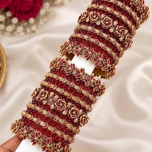 May include: A set of stacked bangles featuring deep red faceted beads and gold-toned metal accents. The design includes alternating bands of plain red beads and decorative gold floral motifs with small white stone inlays, arranged in a repeating pattern.