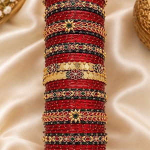 May include: A stack of red bangles featuring gold-toned metal accents, intricate floral motifs, and small green and red inlaid stones. The set includes plain red bangles interspersed with decorative bands that have detailed filigree and patterns.