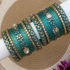 May include: A set of teal-colored bangles featuring gold-toned metal accents and embedded clear stones. The bracelets are arranged in a stack, showcasing intricate floral patterns and small, sparkling embellishments across the textured surfaces.