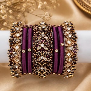 May include: A set of purple silk-wrapped bangles featuring gold-toned metal accents, clear crystal embellishments, and intricate floral patterns. The collection includes plain thread-wrapped bands alongside wider, ornate pieces with embedded stones.