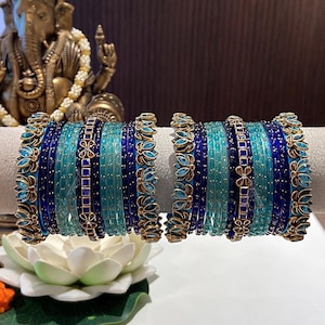 May include: A set of two matching bangle stacks featuring alternating thin bands in shades of royal blue and turquoise. The stacks include textured gold-tone metal bands with blue floral accents and clear crystal embellishments throughout the design.