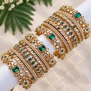May include: A set of gold-toned bangles featuring emerald green square-cut stones and clear white crystals arranged in floral patterns. The bracelets are stacked together, showcasing intricate metalwork and a polished finish on each piece.