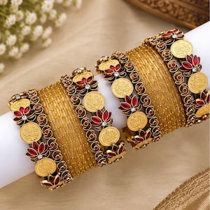 May include: A set of gold-toned bangles featuring thin, textured bands interspersed with decorative elements. These accents include circular gold-coloured coin motifs and floral-shaped pieces inlaid with deep red stones and small clear crystals.