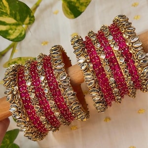 Pode incluir: Um conjunto de pulseiras fúcsia vibrantes com detalhes em metal dourado e pedras transparentes em formato de marquise. As pulseiras estão dispostas em dois grupos numa haste de madeira, exibindo detalhes metálicos intrincados e brilhos.