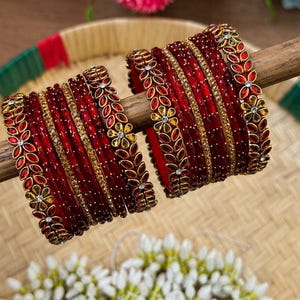 Könnte beinhalten: Ein Set aus roten Armreifen mit goldfarbenen Metallakzenten in einem floralen Blattmuster. Die Armreifen sind mit kleinen klaren Steinen und strukturierten Goldbändern verziert und in zwei Gruppen auf einem Holzstab angeordnet.