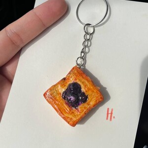 Puede incluir: Un pequeño llavero cuadrado con forma de pastel, con un exterior brillante de color marrón dorado y un relleno morado oscuro. El dije está sujeto a una cadena y un llavero plateados. Una tarjeta blanca con una "H" roja está en el fondo.