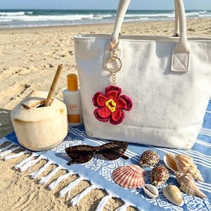 May include: A white canvas tote bag featuring a crocheted flower keychain with dark pink petals and a yellow center. The bag is accompanied by a coconut with a bamboo straw, a bottle of sunscreen, dark brown sunglasses, and several seashells.