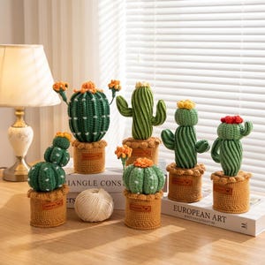 Pode incluir: Uma coleção de cactos de crochê feitos à mão em várias formas e tamanhos. Cada cacto verde tem detalhes amarelos ou vermelhos e está plantado em um vaso de tecido marrom. A cena é montada em uma superfície de madeira.