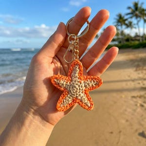 May include: A crocheted starfish keychain featuring a beige center and a bright orange border. The starfish is attached to a gold-toned metal split ring and a lobster clasp by a short chain, creating a decorative accessory for keys or bags.