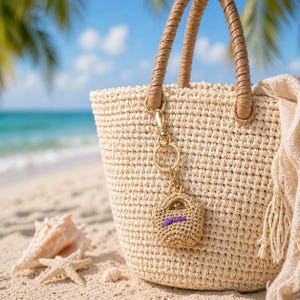 Pode incluir: Uma bolsa tote de palha tecida com alças marrons e um chaveiro dourado. O chaveiro tem um pingente de bolsa tecida em miniatura com um detalhe roxo. A bolsa está em uma praia arenosa com conchas e uma estrela do mar.
