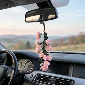 Op de afbeelding: Een hangend auto-accessoire bestaande uit een gehaakte rank met meerdere roze bloemen, gele harten en donkergroene bladeren, opgehangen aan een bruine leren riem aan de achteruitkijkspiegel, wat een decoratieve bloemige uitstraling geeft.