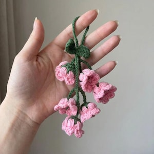 May include: A handmade crochet floral ornament featuring pink bell-shaped flowers and green stems and leaves. The crochet flowers are clustered together, creating a delicate and charming decorative piece. The ornament is held in a hand.