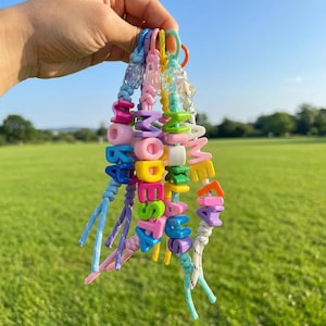 May include: A collection of five colorful beaded keychains featuring plastic letter beads spelling out names including Modesta, Niklaus, and Amelia. Each keychain is constructed from braided cord and includes small flower-shaped accent beads.