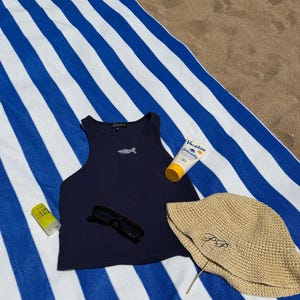 May include: A navy blue tank top with a small embroidered design, sunglasses, and a straw hat with embroidered initials are arranged on a blue and white striped beach towel. A tube of sunscreen and a small bottle of sunscreen are also present.