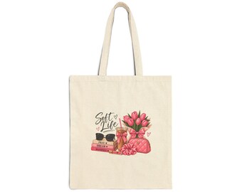 May include: A beige canvas tote bag featuring a printed graphic of pink tulips, a drink with a straw, a quilted pink pouch, a pink scrunchie, black sunglasses, and a stack of pink books labeled Pray, Dream, Manifest with the text Soft Life.