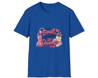 May include: A royal blue short-sleeve t-shirt featuring a graphic print in shades of pink. The design includes the text "Small Steps BIG Dreams" surrounded by illustrations of headphones, a drink, books, a camera, a tote bag, and sneakers.
