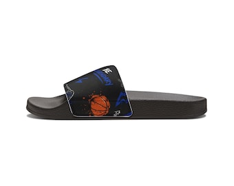 May include: A black slide sandal featuring a patterned strap with an orange basketball graphic, blue lightning bolt icons, and the text BE LEGENDARY printed in blue. The sandal has a contoured footbed and a textured black sole for support.