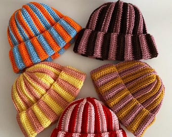 May include: Five crocheted beanies arranged in a circle. One is orange and light blue striped, one is dark brown and mauve striped, one is yellow and light pink striped, one is mustard yellow and mauve striped, and one is red and light pink striped.