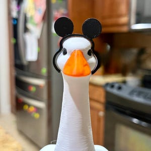 May include: A white 3D printed goose figurine featuring a bright orange beak and black eyes. The goose is wearing a black headband with two circular black ears attached to the top, positioned symmetrically on its head above the eyes.