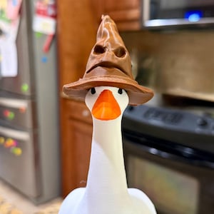 May include: A white goose figurine with an orange beak, wearing a brown, pointed, textured hat with a folded brim and facial features sculpted into the crown. The goose has black eyes and a smooth, matte finish on its neck and body.
