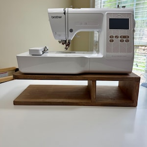 May include: A white Brother sewing machine with a digital display and control buttons. The machine sits on a wooden stand. The stand is made of dark brown wood and has a two-tiered design. The machine is positioned in front of a window.
