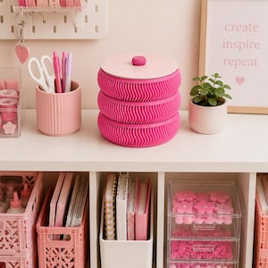 May include: A pink and white storage container with a textured design sits on a white shelf, surrounded by pink stationery. The shelf also holds notebooks, pens, and decorative items, creating a coordinated workspace.