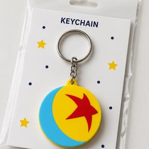 Puede incluir: Un llavero circular con un diseño de pelota amarilla y azul con una estrella roja, unido a una anilla de metal plateado. El llavero está montado sobre una tarjeta de presentación blanca con la palabra KEYCHAIN y estrellas amarillas y puntos azul oscuro.