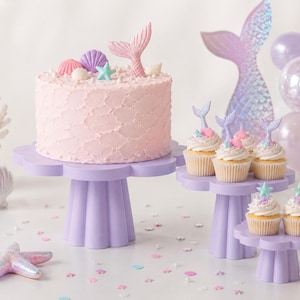 Puede incluir: Un pastel con glaseado rosa claro y varios cupcakes decorados con colas de sirena, conchas y estrellas de mar. Los postres se exhiben en tres soportes para pasteles de color lavanda con bordes festoneados y bases estriadas.