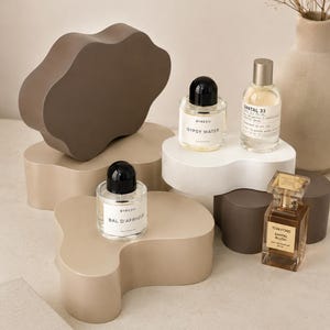 May include: A still-life composition featuring perfume bottles on abstract, neutral-toned pedestals. The bottles include "BYREDO Gypsy Water", "SANTAL 33", "BYREDO Bal d'Afrique", and "TOM FORD Santal Blush". A vase with dried flowers and a book are also present.