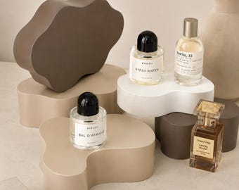 May include: Three glass perfume bottles labelled Byredo Gypsy Water, Byredo Bal d'Afrique, and Tom Ford Santal Blush, along with a Le Labo Santal 33 bottle, displayed on various wavy-shaped beige, brown, and white pedestals near a book.