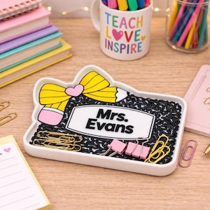 Puede incluir: Un organizador de escritorio blanco con forma de lápiz con un lazo amarillo y una goma de borrar rosa. El organizador está lleno de gomas elásticas negras, clips dorados y rosas, y tiene el nombre "Mrs. Evans". Otros útiles de escritorio son visibles.