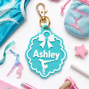 Puede incluir: Un llavero de silicona color verde azulado con borde festoneado, que presenta un lazo blanco en la parte superior, el nombre Ashley en letras blancas y la silueta blanca de una persona haciendo el pino, unido a un mosquetón dorado.