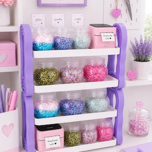 May include: A four-tiered white storage rack with purple side supports holding clear jars filled with colorful glitter, sprinkles, and confetti. Two pink label makers are present, each printing a white label that reads Handmade with heart.