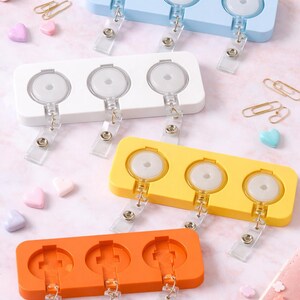 Puede incluir: Una colección de portatarjetas retráctiles en varios colores: azul claro, blanco, amarillo y naranja. Cada soporte tiene tres carretes circulares con clips de plástico transparente para sujetar tarjetas de identificación o insignias. El fondo incluye artículos de papelería y elementos decorativos.