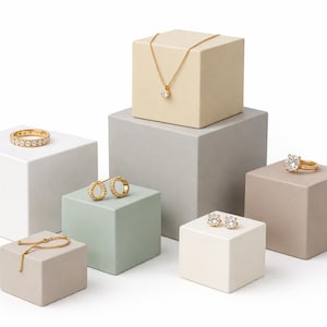 Puede incluir: Una variedad de joyas de oro, incluyendo anillos, pendientes, un collar y una pulsera, exhibidas sobre bloques cuadrados de varios colores neutros. Las joyas presentan piedras preciosas transparentes y están sobre un fondo blanco.