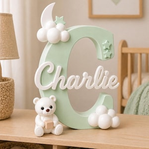 Puede incluir: Una letra "C" verde menta con el nombre "Charlie" en escritura blanca, adornada con nubes blancas, estrellas y una luna creciente. Un pequeño oso de peluche blanco sosteniendo un bloque está frente a la letra. La decoración está sobre una superficie de madera.