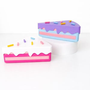 May include: Two colorful cake slice-shaped boxes. One has white frosting with pink and yellow sprinkles, the other has purple frosting with blue and pink sprinkles. Both have a layered design with pink and blue sections.